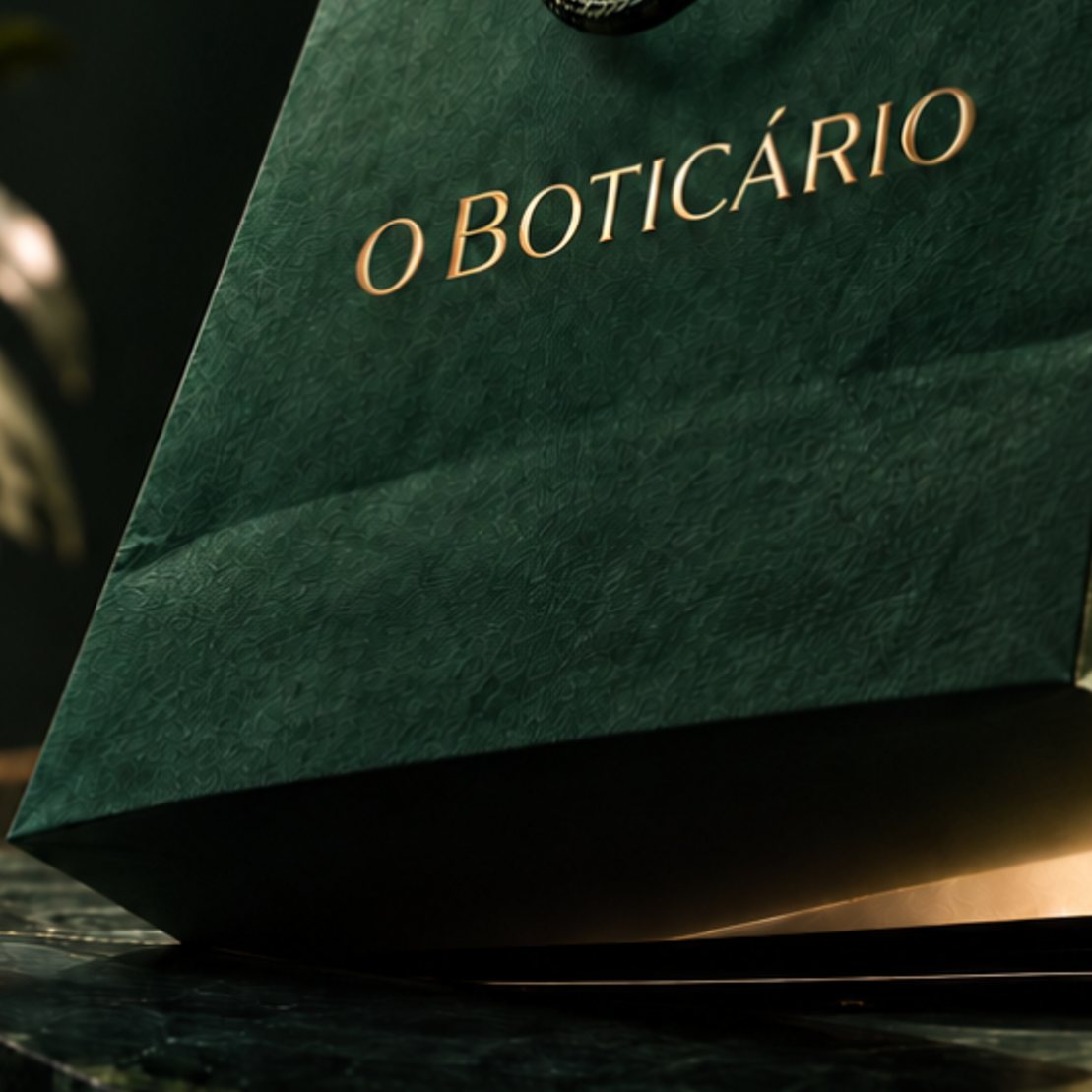 Sacola verde de O Boticário saindo da tela de um celular, sobre uma bancada de mármore escuro